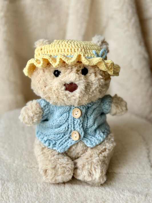 Jellycat Bart Junior wearing a handmade sunny yellow crochet bonnet and baby blue cable knit cardigan on a soft cream blanket.