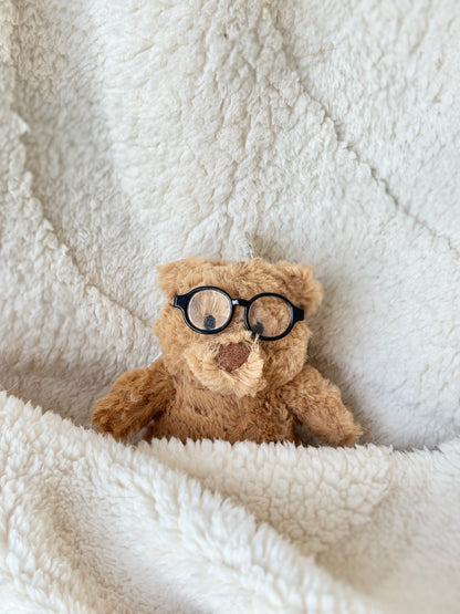 Mini teddy bear Jellycat bag charm tucked in a fluffy cream blanket wearing tiny black round glasses.