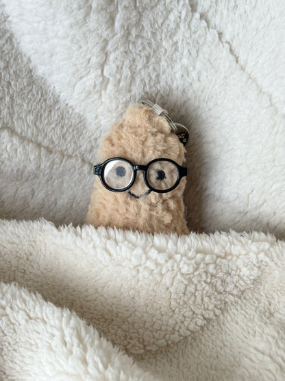 Jellycat peanut bag charm smiling under a blanket while wearing tiny black round glasses.