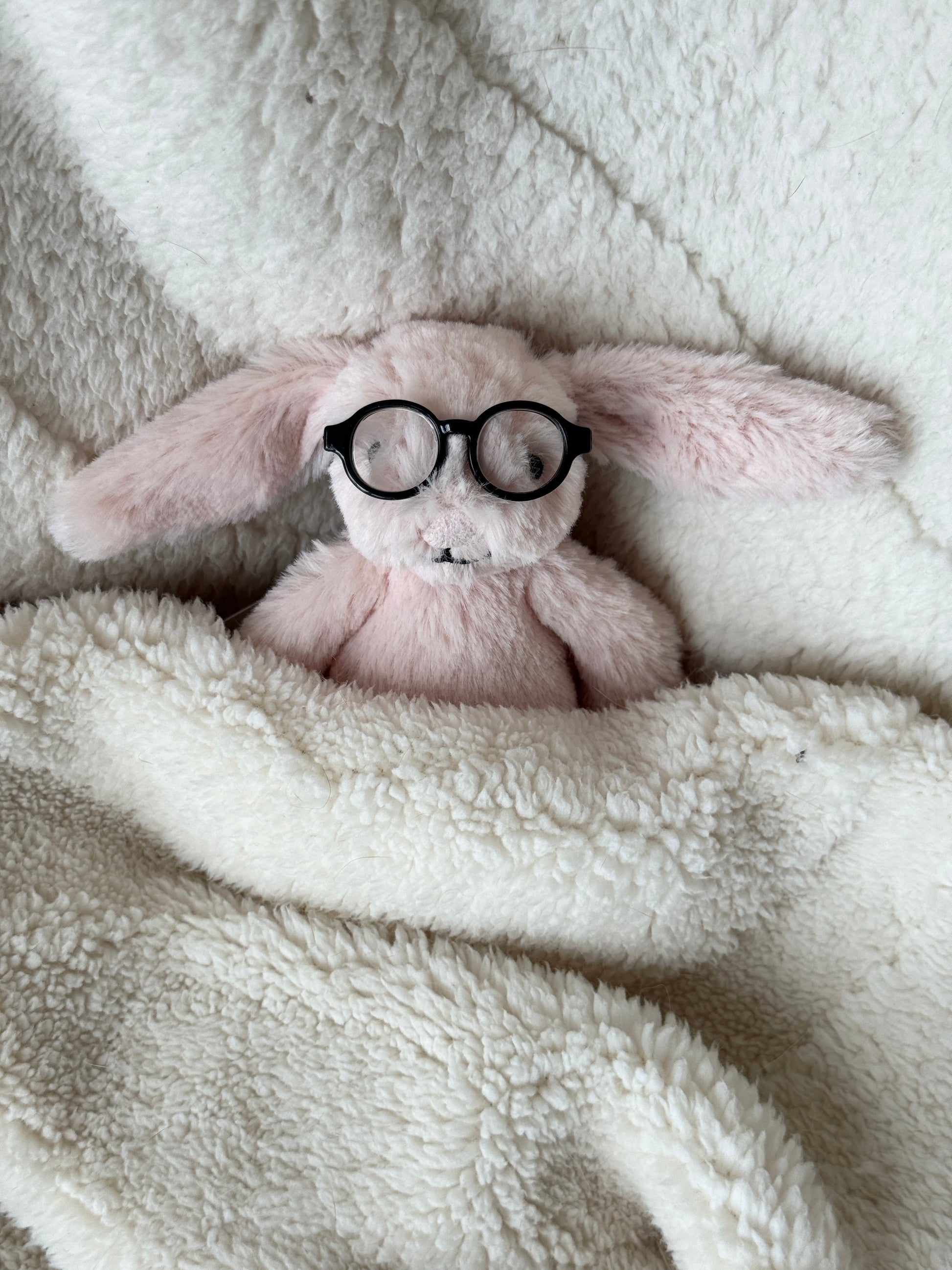 Soft pink bunny Jellycat bag charm resting in a blanket with tiny black round glasses.