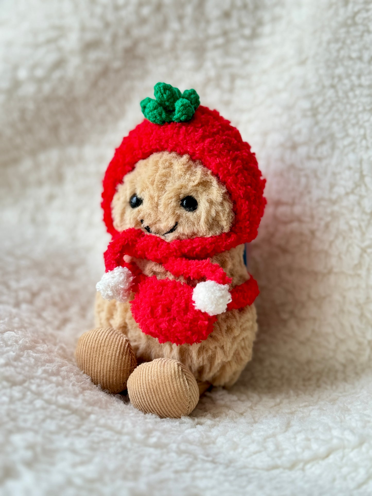 Jellycat Peanut wearing red crochet tomato bonnet and matching scarf with white pompoms, three-quarter angle view on cream background