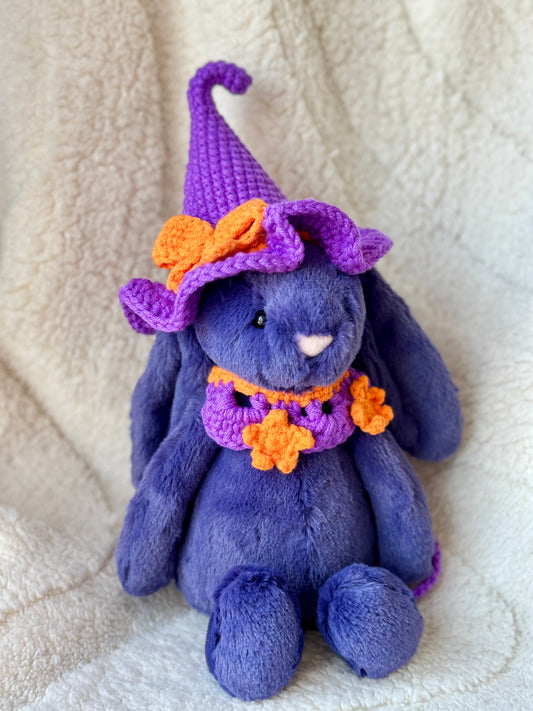 Purple Jellycat bunny wearing a violet crochet witch hat with orange bow and matching star collar.