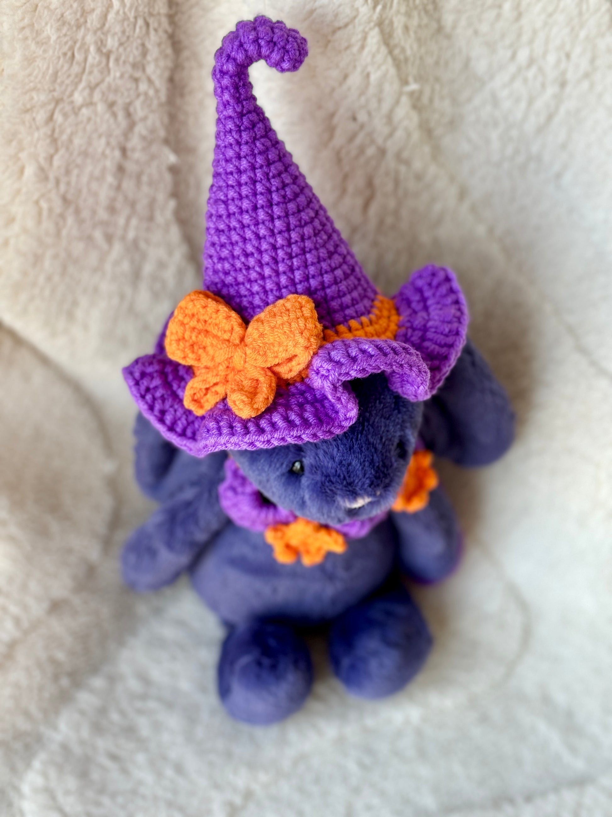 Upper view of Purple Jellycat bunny wearing a violet crochet witch hat with orange bow and matching star collar.