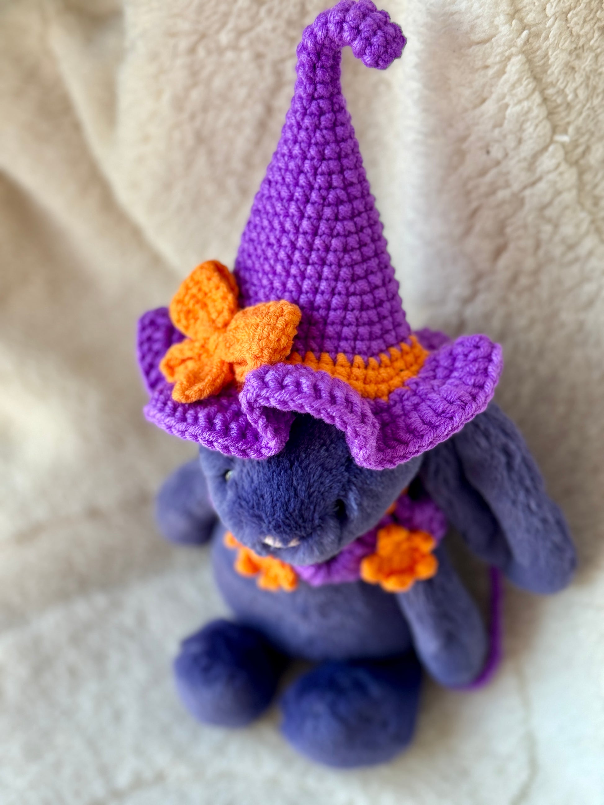 Upper view of Purple Jellycat bunny wearing a violet crochet witch hat with orange bow and matching star collar.

