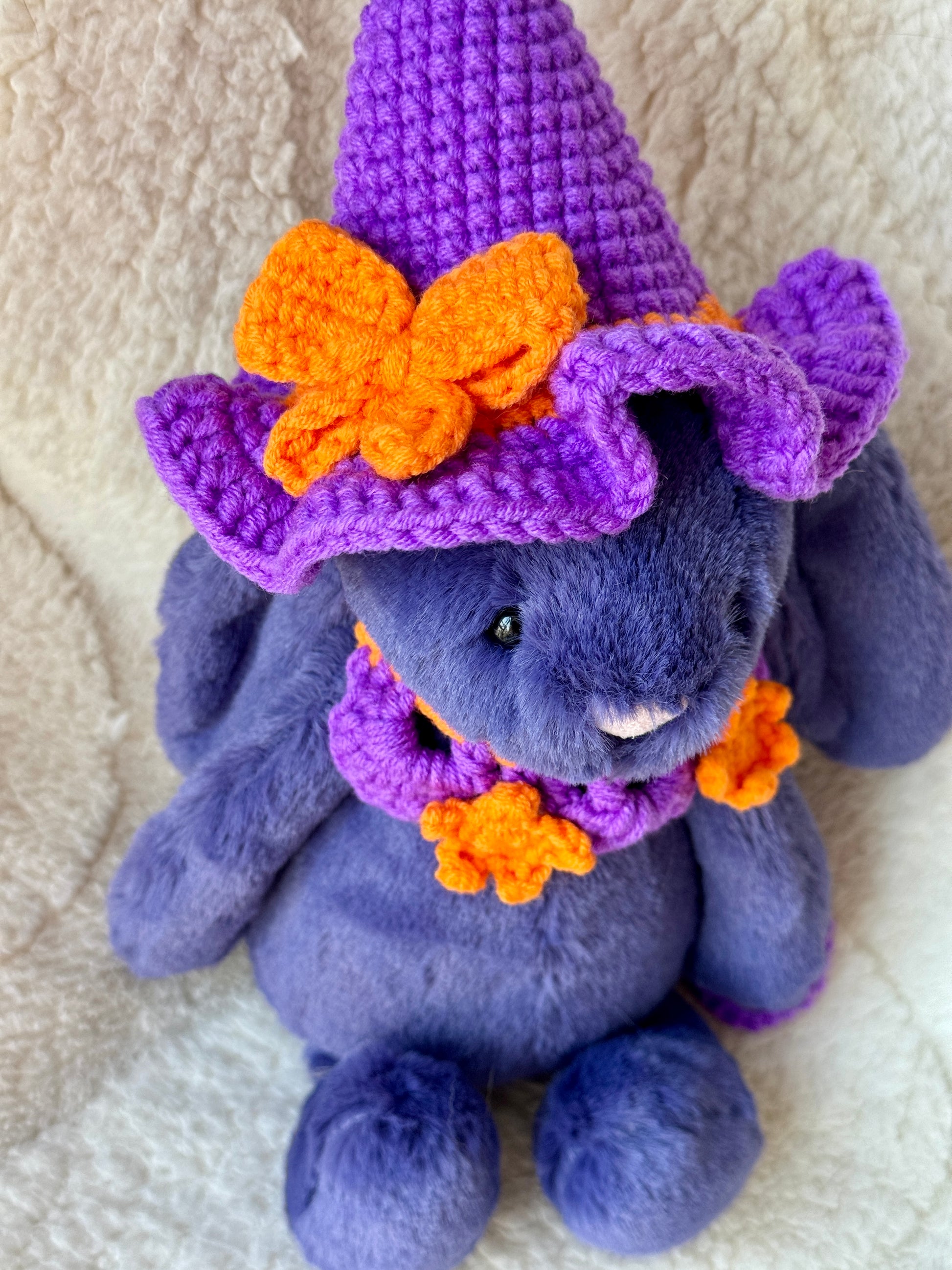 Close view of Purple Jellycat bunny wearing a violet crochet witch hat with orange bow and matching star collar.